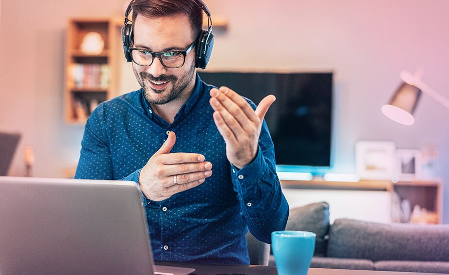 Bespectacled, bearded man in home office, wearing headphones, smiling and gesturing during an online meeting