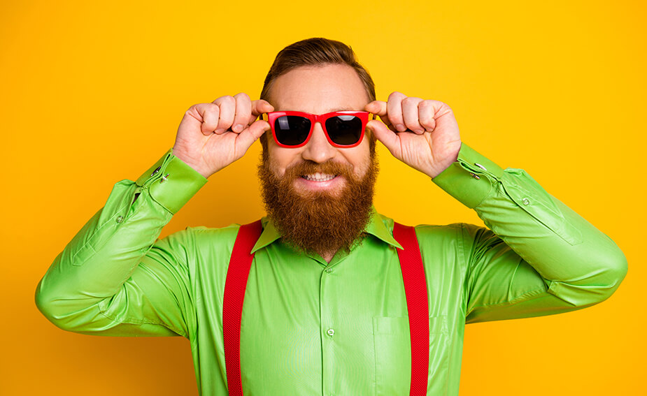 Bearded man wearing green shirt, red suspenders and sunglasses, smiling and standing in front of orange background